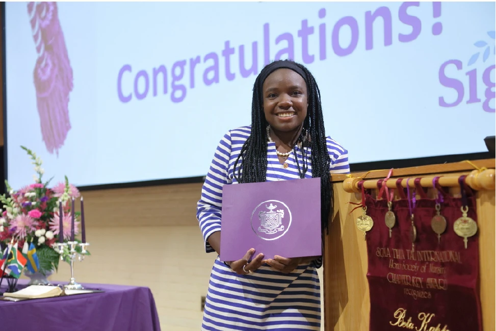 Black woman standing next to a podium holding an award with Congratulations projected on the screen behind her.