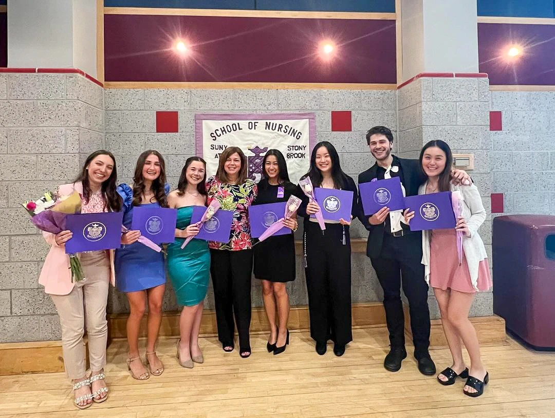 A group on people standing and smiling holding Sigma Theta Tau folders with a banner behind saying School of Nursing, SUNY, Stony Brook.